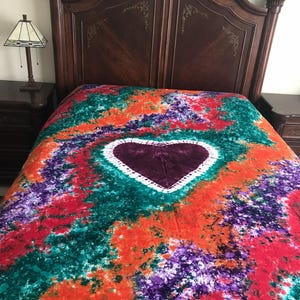 May include: A colorful tie-dye bedspread with a large heart design in the center. The bedspread features a vibrant mix of red, orange, green, and purple hues. The heart is a deep burgundy color with a white border.