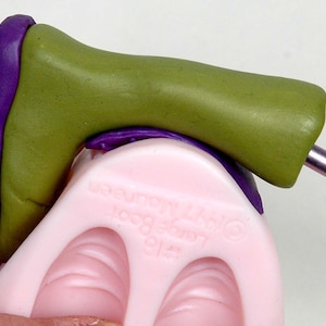 Puede incluir: Un molde de silicona verde y morado con forma de bota para hacer miniaturas de arcilla polimérica. El molde está hecho de silicona y tiene una superficie lisa. El molde se está utilizando para crear una bota en miniatura.