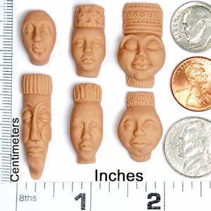 Puede incluir: Siete pequeñas figuritas de terracota pintadas a mano que representan rostros humanos con diferentes expresiones. Las figuritas miden aproximadamente 2,5 cm de alto y están dispuestas en fila sobre una superficie blanca.