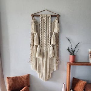 May include: A large macrame wall hanging with a wooden dowel at the top. The macrame is made with natural colored cotton cord and features a variety of knots and tassels. The wall hanging is hanging on a white wall and there is a wooden table with a plant and a vase on it.