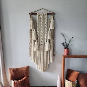 May include: A beige macrame wall hanging with a wooden dowel at the top and a fringe of tassels at the bottom. The wall hanging is hanging on a white wall with a brown rug on the floor.