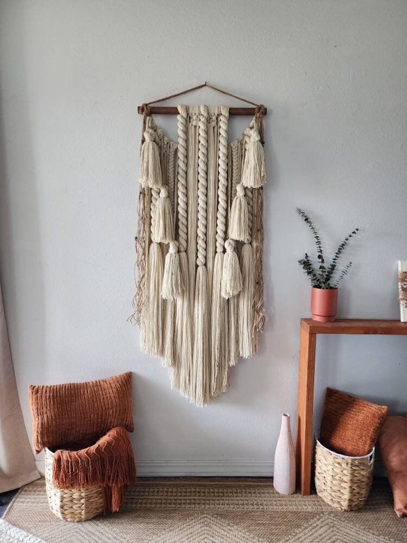 May include: A large macrame wall hanging with a wooden dowel at the top. The macrame is made with natural colored cotton cord and features a variety of knots and tassels. The wall hanging is hanging on a white wall and there is a wooden table with a plant and a vase on it.