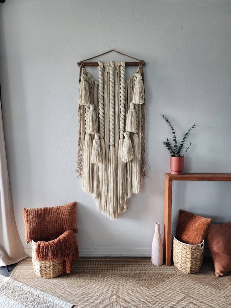 May include: A beige macrame wall hanging with a wooden dowel at the top and a fringe of tassels at the bottom. The wall hanging is hanging on a white wall with a brown rug on the floor.