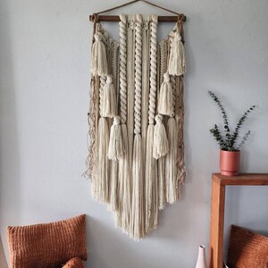 May include: A large, beige macrame wall hanging with a wooden dowel at the top. The hanging features a variety of knots and textures, including tassels and fringe. The hanging is displayed on a white wall with a brown wooden table and a potted plant to the right.