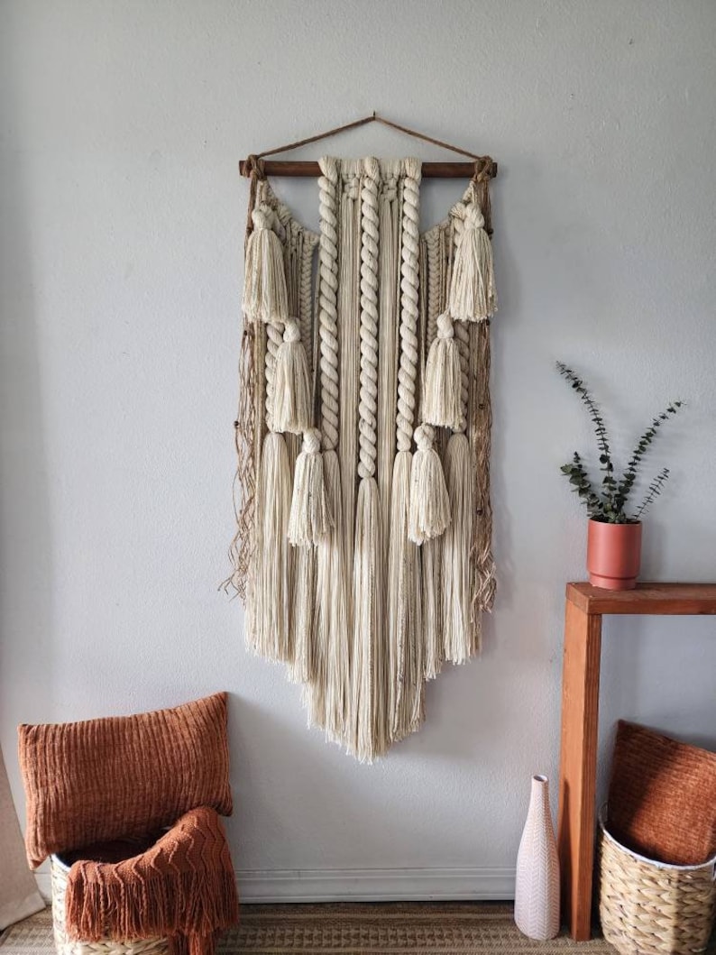 May include: A large, beige macrame wall hanging with a wooden dowel at the top. The hanging features a variety of knots and textures, including tassels and fringe. The hanging is displayed on a white wall with a brown wooden table and a potted plant to the right.