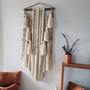 May include: A large, cream-colored macrame wall hanging with tassels and a wooden dowel at the top. The hanging is displayed on a white wall with a small potted plant and a wooden table below.
