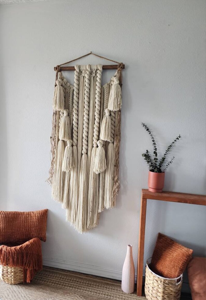 May include: A large, cream-colored macrame wall hanging with tassels and a wooden dowel at the top. The hanging is displayed on a white wall with a small potted plant and a wooden table below.
