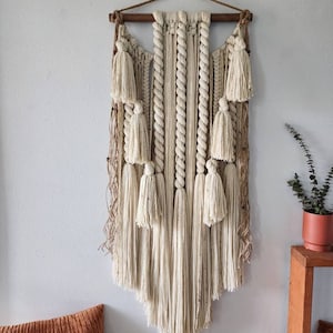 May include: A large, off-white macrame wall hanging with a wooden dowel at the top. The hanging features a variety of knots and textures, including rope, yarn, and fringe. The fringe is long and flowing, creating a soft and airy look.