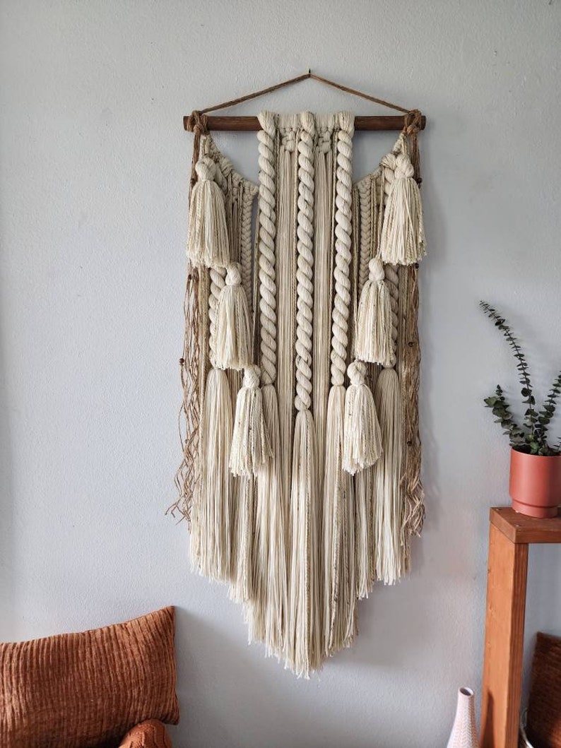 May include: A large, off-white macrame wall hanging with a wooden dowel at the top. The hanging features a variety of knots and textures, including rope, yarn, and fringe. The fringe is long and flowing, creating a soft and airy look.
