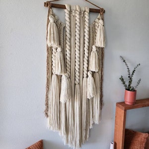 May include: A large, off-white macrame wall hanging with a wooden dowel at the top. The macrame features a variety of knots and textures, including tassels and fringe. The wall hanging is hanging on a white wall.