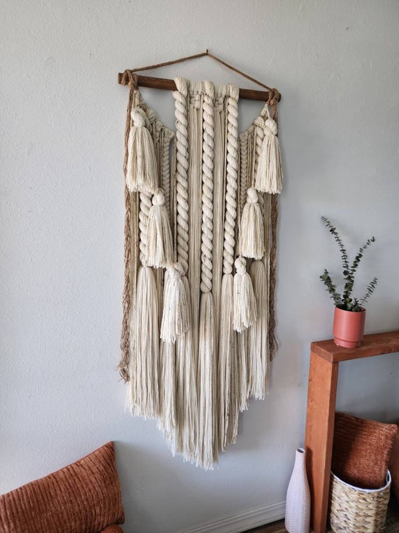 May include: A large, off-white macrame wall hanging with a wooden dowel at the top. The macrame features a variety of knots and textures, including tassels and fringe. The wall hanging is hanging on a white wall.