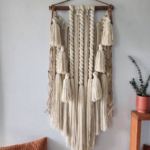 May include: A large, off-white macrame wall hanging with a wooden dowel at the top. The hanging features a variety of knots and textures, including rope, yarn, and fringe. The fringe is long and flowing, creating a soft and airy look.