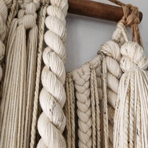 May include: A white macrame wall hanging with a wooden dowel at the top. The macrame is made with a variety of knots and textures, including braids, twists, and tassels.