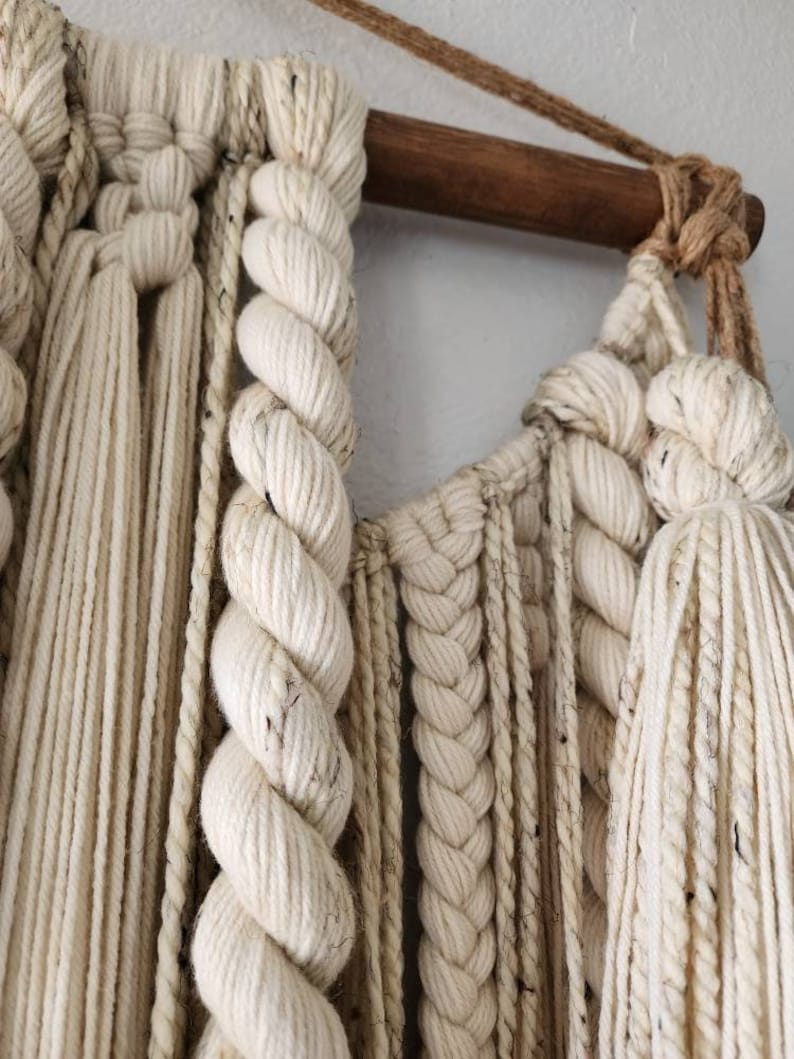 May include: A white macrame wall hanging with a wooden dowel at the top. The macrame is made with a variety of knots and textures, including braids, twists, and tassels.