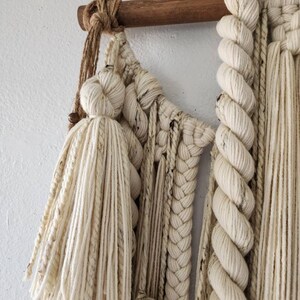 May include: A white macrame wall hanging with a wooden dowel at the top. The macrame is made with a variety of knots and textures, including tassels and braids.