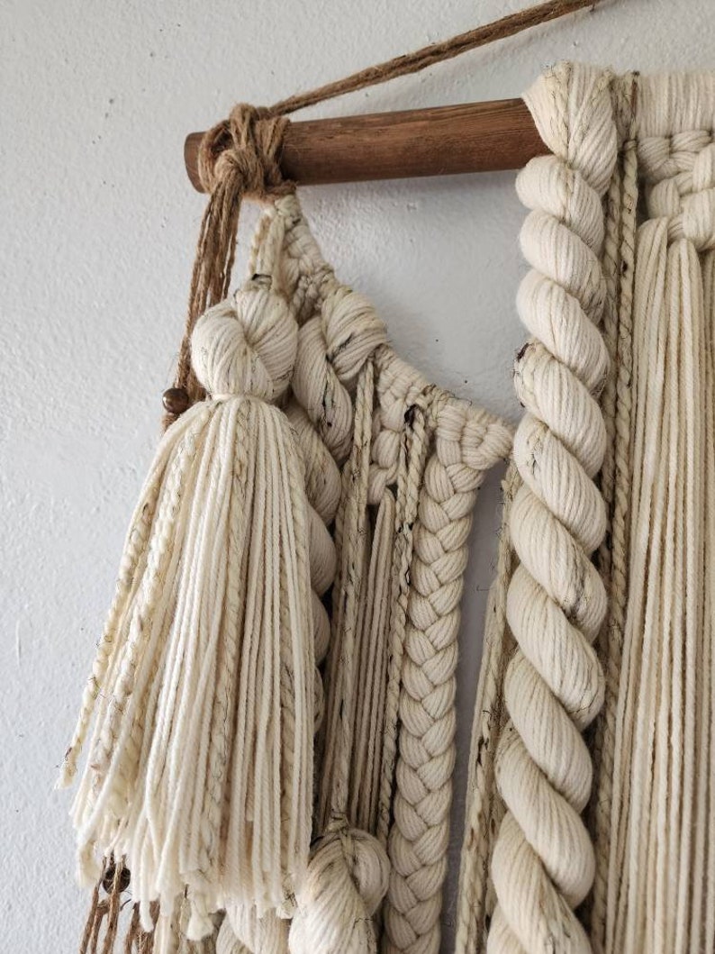 May include: A white macrame wall hanging with a wooden dowel at the top. The macrame is made with a variety of knots and textures, including tassels and braids.