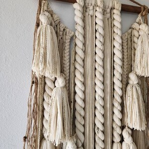 May include: A white macrame wall hanging with a wooden dowel at the top. The hanging features a variety of knots and textures, including tassels and braids.
