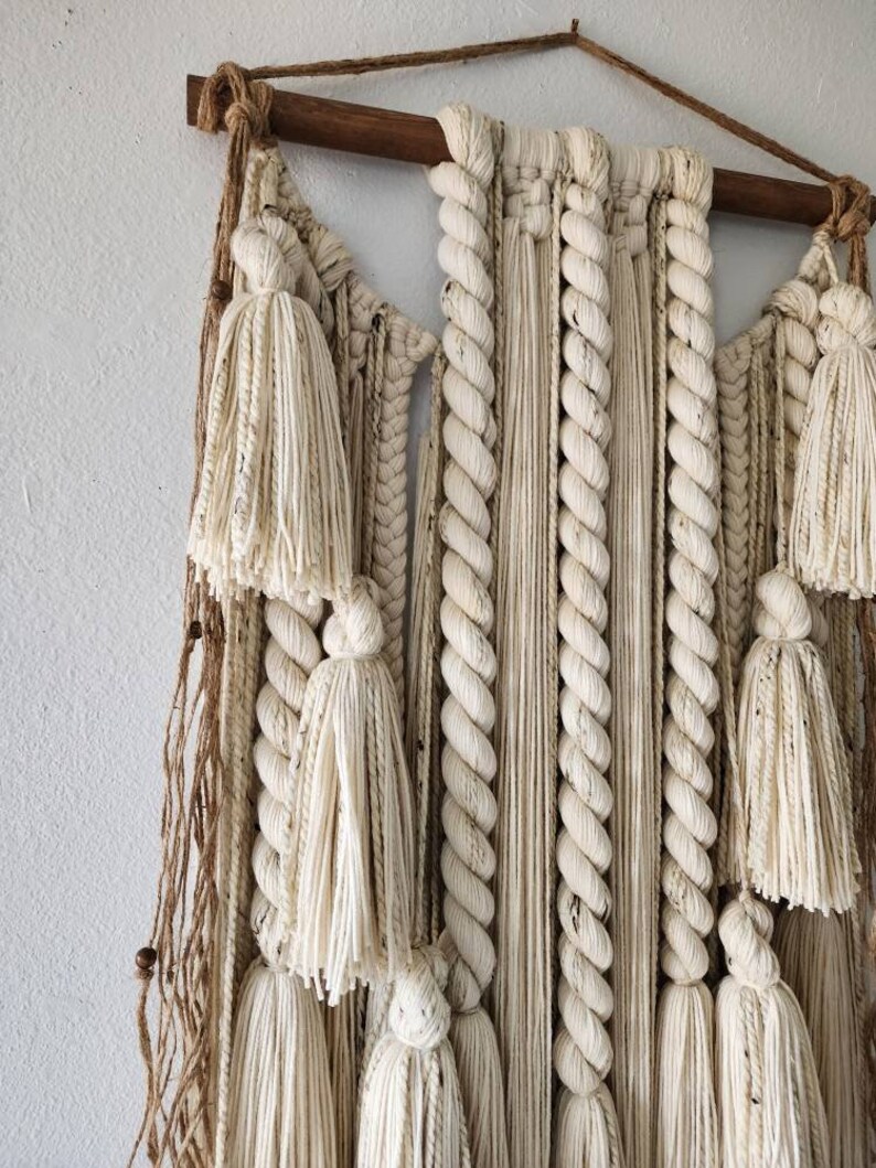 May include: A white macrame wall hanging with a wooden dowel at the top. The hanging features a variety of knots and textures, including tassels and braids.