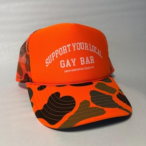 May include: Bright orange trucker hat with the text "SUPPORT YOUR LOCAL GAY BAR" in white. The hat features a camouflage pattern on the brim and mesh sides. The text "RNBW QUEER MUSIC COLLECTIVE" is printed below the main text.