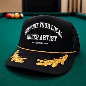 May include: A black trucker hat featuring a mesh back and a gold rope trim across the brim. The front displays white text reading SUPPORT YOUR LOCAL QUEER ARTIST and GIRLCRUSH SOCIAL HOUSE, with gold embroidered leaf designs on the brim.