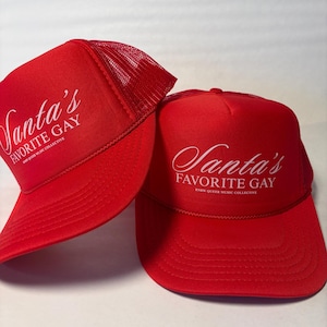 May include: Two red trucker hats with white text that reads "Santa's Favorite Gay" in a script font. The hats have a mesh back and a curved brim. The text "KNOW QUEER MUSIC COLLECTIVE" is below the main text.