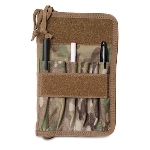 May include: A camouflage-patterned notebook cover with multiple pockets and a zipper closure. The cover has a brown velcro strap and includes a black pen, a black marker, and a white pen.