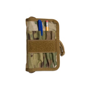 May include: A camouflage-patterned notebook cover with a brown border and a Velcro fastening. A blue pen and a pencil are visible in the pockets. The notebook has a zip closure and a drawstring.