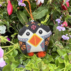 Puede incluir: Un adorno de tela acolchado hecho a mano con forma de pingüino, con bordado de punto de cruz en negro, blanco y naranja. Tiene un pico naranja brillante, dos pequeños ojos negros y un lazo para colgar trenzado en amarillo y rojo con borlas naranjas.