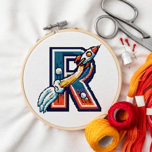 Op de afbeelding: Een kruissteek-borduurring met een kleurrijk gepixeld ontwerp van de letter "R" met een raket erin. De ring ligt op een witte stoffen achtergrond met kleurrijke borduurgarenrollen.