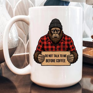 May include: White ceramic coffee mug featuring a Bigfoot-like creature wearing a black beanie and a red and black plaid shirt. The creature holds a wooden sign that reads "DO NOT TALK TO ME BEFORE COFFEE."