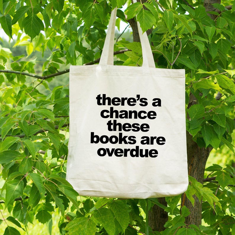 A white tote bag with the text "there's a chance these books are overdue" printed on it, hanging from a tree branch.
