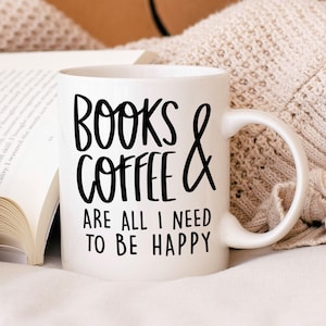 A white mug with black text that reads "BOOKS & COFFEE ARE ALL I NEED TO BE HAPPY" is placed on a white surface, with an open book and a cozy blanket nearby.