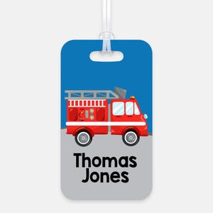May include: A rectangular luggage tag with a clear plastic loop at the top. The design features a red fire truck on a blue and gray background. The text "Thomas Jones" is printed in bold black letters at the bottom of the tag.