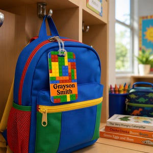 May include: A blue backpack with green and yellow accents features a rectangular luggage tag attached to the top handle. The tag displays a colorful interlocking brick pattern and the printed name Grayson Smith in bold black capital letters.