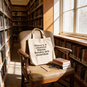 A beige tote bag with the text "There's a chance these books are overdue" sits on a wooden chair in a library, with a stack of books and a pair of glasses nearby.
