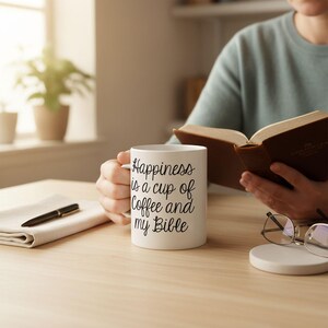 A person is sitting at a desk, reading a book and holding a mug with the text "Happiness is a cup of coffee and my Bible" printed on it.