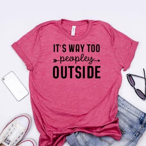 A pink t-shirt with the text "IT'S WAY TOO PEOPLEY OUTSIDE" printed on it, along with a pair of jeans and a pair of sunglasses.