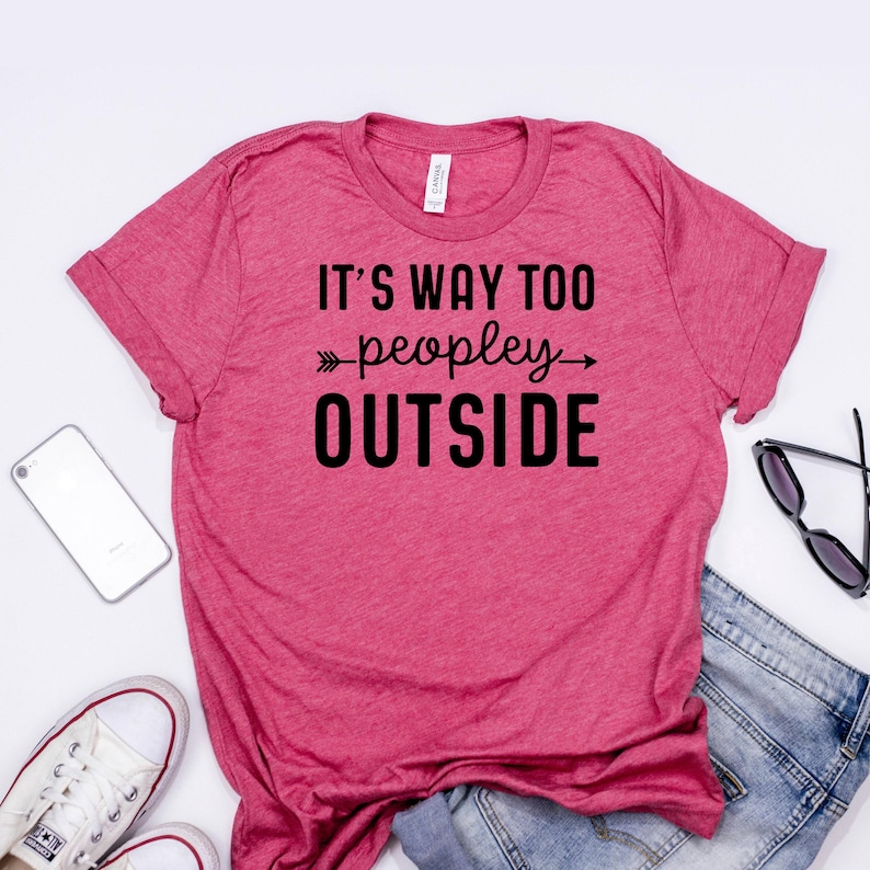 A pink t-shirt with the text "IT'S WAY TOO PEOPLEY OUTSIDE" printed on it, along with a pair of jeans and a pair of sunglasses.