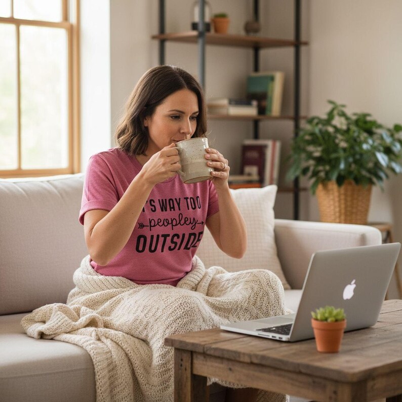A woman sits on a couch, wrapped in a blanket, sipping a coffee and using a laptop.