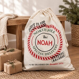 May include: A cream-coloured canvas drawstring sack featuring a red and green circular North Pole stamp design. The text reads FIRST CLASS, NORTH POLE to: NOAH, SPECIAL DELIVERY, EXPRESS, and TO BE DELIVERED ON december 25TH in black and red ink.