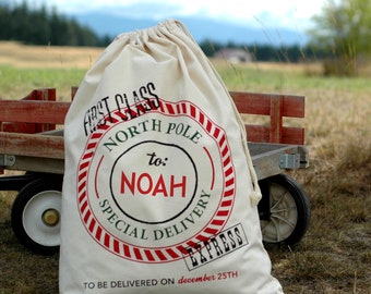 May include: A cream-colored canvas drawstring sack featuring a red and white striped circular stamp design. The text reads FIRST CLASS, NORTH POLE, to: NOAH, SPECIAL DELIVERY, EXPRESS, and TO BE DELIVERED ON december 25TH in black and red ink.