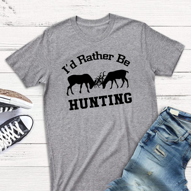 A gray t-shirt with the text "I'd Rather Be HUNTING" printed on it, along with two silhouetted deer figures. The t-shirt is laid out on a wooden surface, and there are a pair of jeans and a pair of sneakers visible in the background.