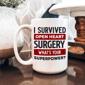 May include: White ceramic mug with a large handle. The mug features the text "I SURVIVED OPEN HEART SURGERY WHAT'S YOUR SUPERPOWER?" in black and red lettering. The mug is sitting on a brown surface.