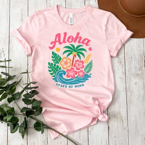 A pink t-shirt with a colorful graphic design of a palm tree, flowers, and waves, along with the text "Aloha State of Mind" printed on it. The t-shirt is placed on a wooden surface, and there is a brown hat nearby.