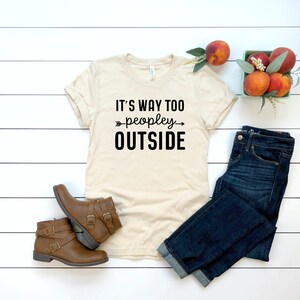 a casual outfit consisting of a white t-shirt with the text "IT'S WAY TOO PEOPLEY OUTSIDE", a pair of blue jeans, brown boots, and a small basket of peaches.