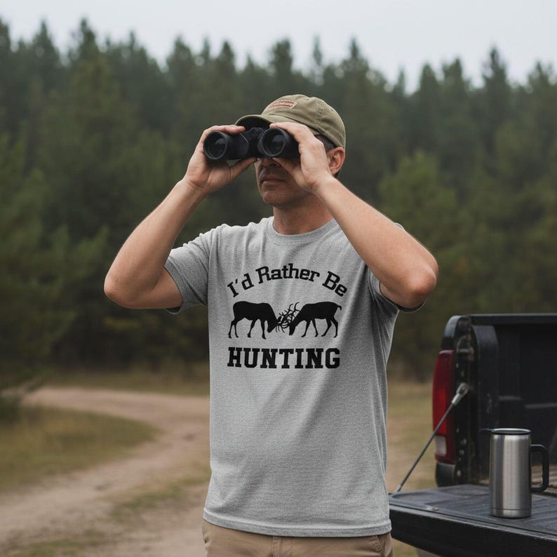 A man wearing a t-shirt with the text "I'D Rather Be HUNTING" is standing in front of a truck, looking through binoculars.