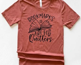 May include: A brick-red t-shirt with a crew neck. The shirt features a black graphic of an open book with the text "Bookmarks are for Quitters" in a handwritten style. The design includes stars above and below the book.