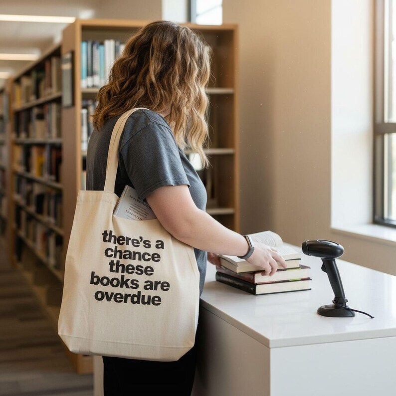 A woman is standing in a library, holding a white tote bag with the text "There's a chance these books are overdue" printed on it. She is looking at a stack of books on a table in front of her.