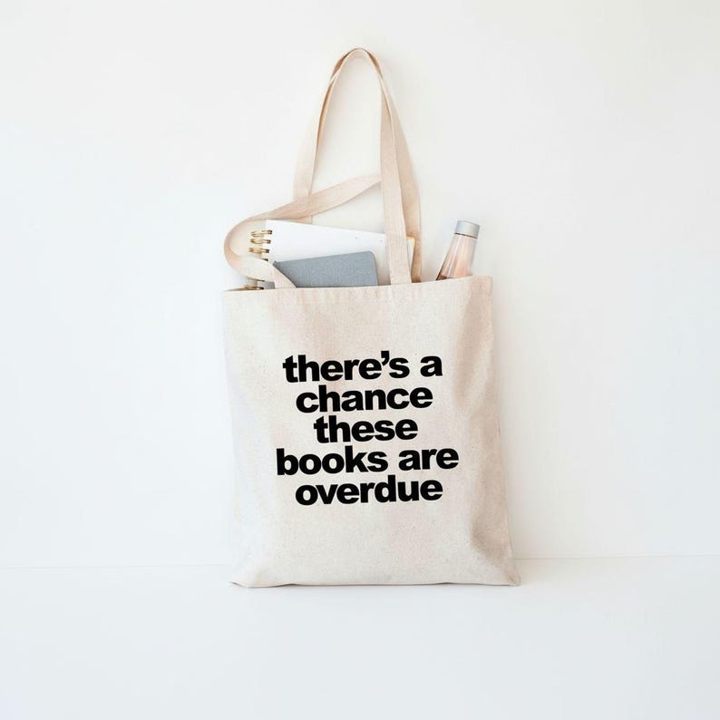 A beige tote bag with the text "there's a chance these books are overdue" printed on it, along with a few books and a bottle inside the bag.