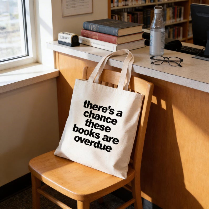 A white tote bag with the text "there's a chance these books are overdue" printed on it, sitting on a wooden chair in a library or bookstore setting.